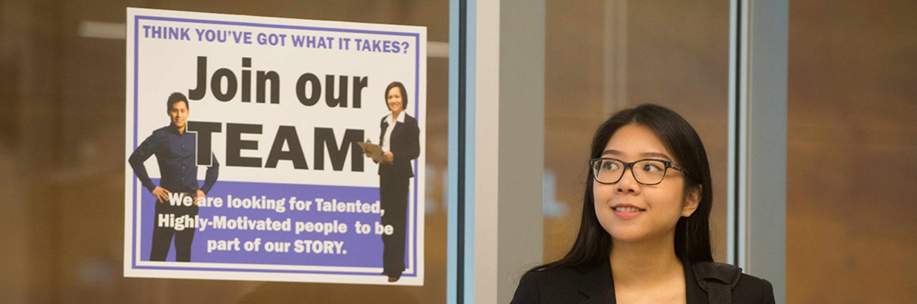 Person wearing business attire standing in the doorway with a "Join our team" sign posted